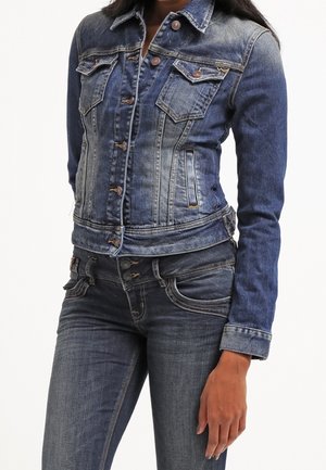 Femme portant une veste en jean bleu ajustée avec fermeture par boutons et un jean en denim bleu assorti avec coutures visibles et braguette à boutons.