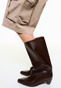 Cizme din piele maro, până la genunchi, cu vârf ascuțit și toc mic, asortate cu pantaloni scurți bej, până la genunchi, care au buzunare laterale.