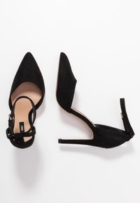 Černé semišové vysoké podpatky mají špičatou špičku, v�ýřezy na bocích a tenký kotníkový pásek se zlatou sponou. Design s podpatkem stiletto zvýrazňuje eleganci.