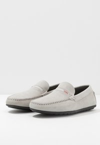 Mocassins en daim gris avec des détails cousus, une semelle en caoutchouc plate et un petit logo rouge sur le côté. Texture lisse et forme arrondie.