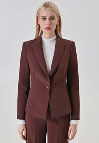 Blazer color granata con design aderente, chiusura con un solo bottone caratterizzata da un accento circolare, rever affilati e dettagli sottili delle cuciture.