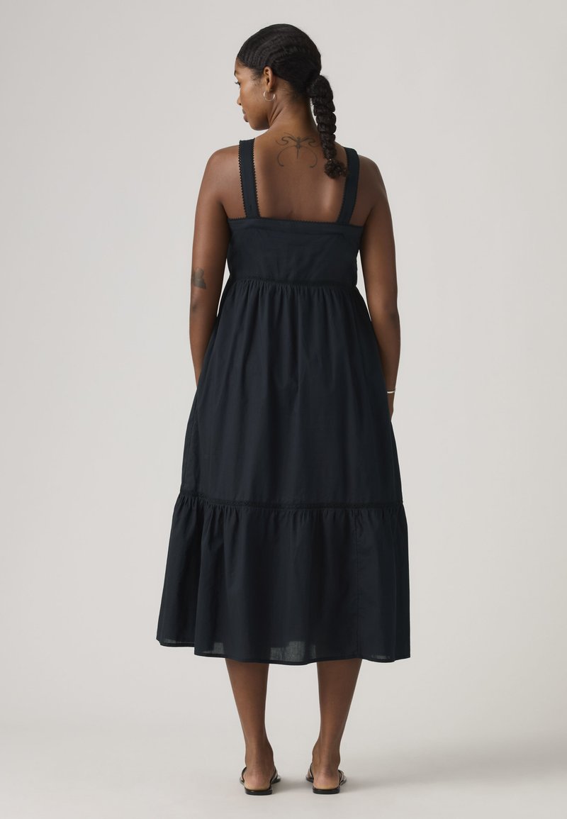Femme aux cheveux tressés portant une robe midi noire sans manches avec des larges bretelles et un ourlet à volants, debout de dos face à la caméra.