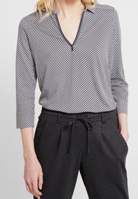 Blusa a scacchi bianca e nera con scollatura a V e maniche a tre quarti, abbinata a pantaloni a scacchi grigi scuri con vita regolabile.