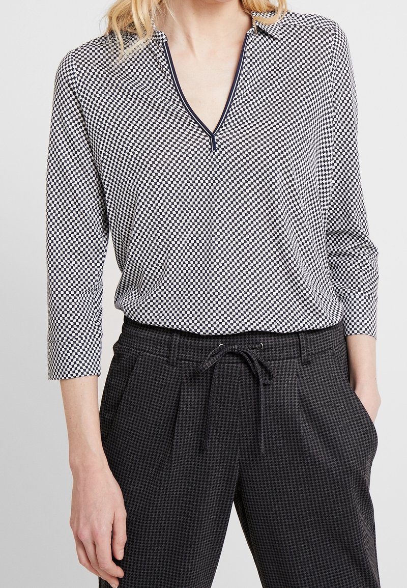 Blusa a scacchi bianca e nera con scollatura a V e maniche a tre quarti, abbinata a pantaloni a scacchi grigi scuri con vita regolabile.