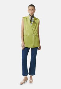 Jasnozielona bezrękawnik z podwójnymi guzikami, w połączeniu z rozszerzającymi się niebieskimi dżinsami i białymi sandałami na obcasie. Akcent w postaci wzorzystego jedwabnego szala.