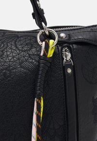Sac en cuir noir texturé avec fermeture éclair argentée et détail anneau, avec des cordons colorés enroulés attachés à l’anneau près de la poignée.