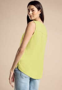 Top senza maniche verde lime con orlo arrotondato, realizzato in tessuto morbido, caratterizzato da una sottile cucitura sul retro e fessure laterali, abbinato a jeans azzurri chiari.