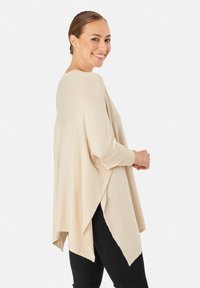 Lächelnde Frau, leicht zur Seite gedreht, trägt einen lockeren beigen Poncho mit seitlichen Schlitzen über einer schwarzen Hose, vor einem schlichten weißen Hintergrund.