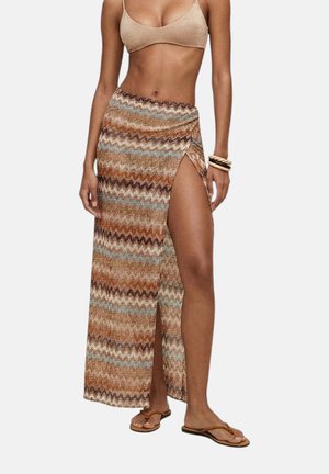 Modella che indossa una gonna maxi multicolore con motivo a zigzag e una spacco alto sulla coscia, abbinata a un top corto beige e sandali.