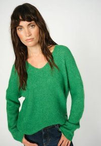 Groene gebreide trui met een losse pasvorm, V-hals en lange mouwen. Zachte textuur met een lichte glans. Gepaarde met donkere spijkerbroek.