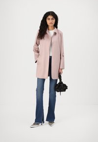 Lys rosa, knelang frakk med en jevn tekstur, som har detaljer på krage og mansjetter. Kombinert med utsvingte blå jeans og metalliske sko.