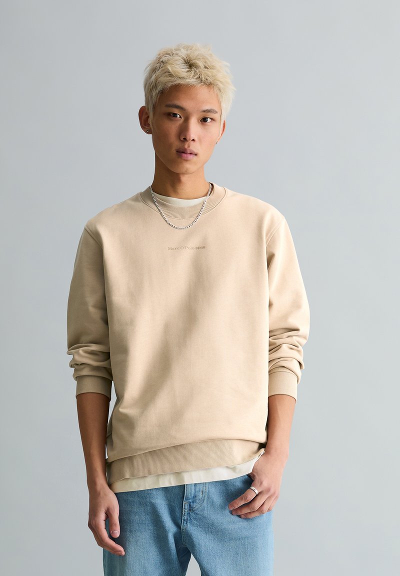 Νέος άνδρας με κοντά ξανθά μαλλιά, ντυμένος με μπεζ φούτερ, ασημένια κολιέ, ανοιχτό μπλε τζιν και στέκεται σε απλό γκρι φόντο.