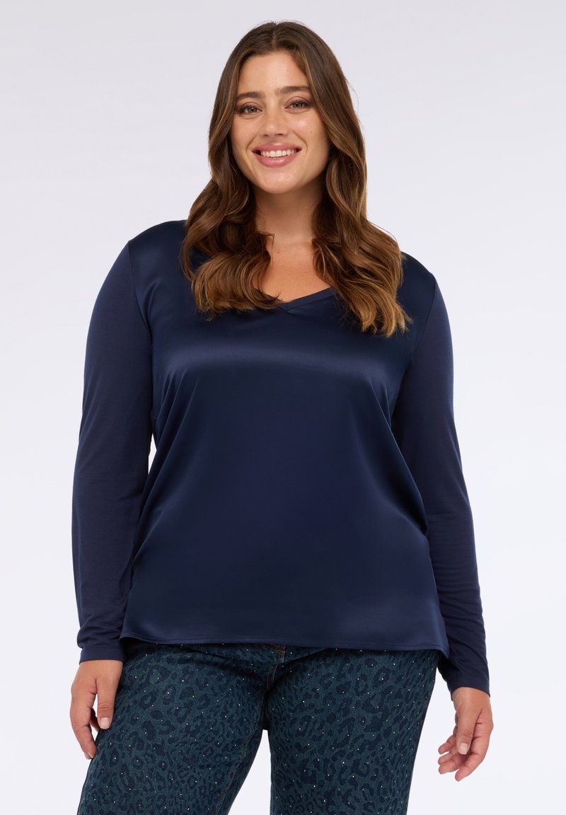 Navyblaues Langarm-V-Ausschnitt-Oberteil aus einem glatten, satinähnlichen Material, kombiniert mit dunklen gemusterten Jeans, die ein dezentes Leopardenmuster aufweisen.