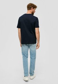 Námořnické bavlněné tričko s krátkým rukávem, v kombinaci se světle modrými džíny. Model v bílých teniskách, pohled zezadu.