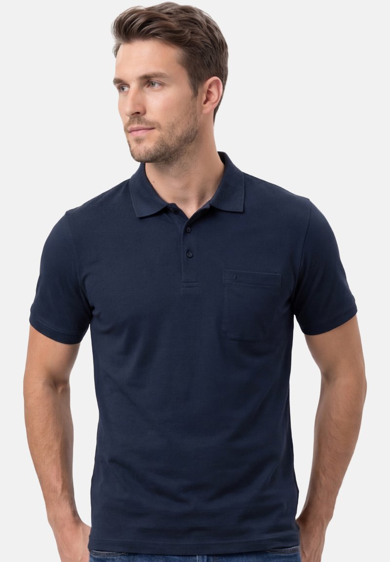 Homme portant un polo bleu marine à manches courtes avec une poche poitrine, regardant vers la gauche sur un fond blanc.