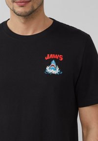 Schwarzes Baumwoll-T-Shirt mit Rundhalsausschnitt, das roten "JAWS"-Text und ein gesticktes Haifischdesign in Blau und Weiß zeigt. Mit kurzen Ärmeln und lässigem Schnitt.