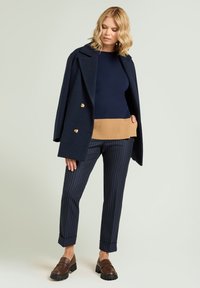 Abrigo azul marino cruzado, suéter de canalé azul oscuro con dobladillo beige, pantalones a rayas y mocasines marrones con suelas gruesas.