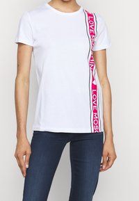 Vit t-shirt med rundad halsringning som har rosa grafiska text och svarta accenter längs ena sidan. Korta ärmar, mjukt bomullsmaterial.