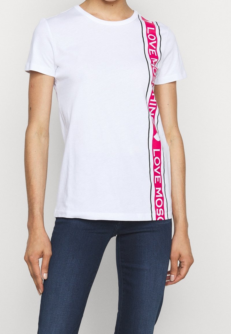 Vit t-shirt med rundad halsringning som har rosa grafiska text och svarta accenter längs ena sidan. Korta ärmar, mjukt bomullsmaterial.