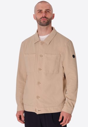 Uomo con barba corta che indossa una giacca beige con bottoni, due tasche sul petto e una toppa circolare nera sulla manica sinistra, sfondo neutro.