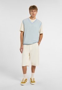 Světle modrá, texturovaná vesta přes krémovou košili; v kombinaci s krémovými šortkami a hořčicovými teniskami. Neformální, vrstvený outfit s moderním střihem.