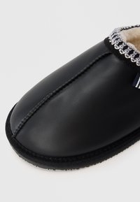 Czarne klapki wsuwane z zaokrąglonym noskiem, górna część z gładkiego materiału przypominającego skórę, szare przeszycia i teksturowana gumowa podeszwa. Widoczna miękka podszewka wewnętrzna.