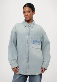 Gaiši zila džinsu krekls ar pogu priekšā, norādītu apkakli un priekšējo kabatu, uz kuras ir uzraksts "MOSCHINO JEANS" zilā krāsā.