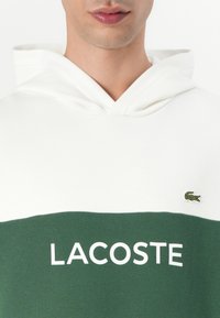 Valge ja rohelise kapuutsiga sviiter, millel on silmapaistev valge "LACOSTE" tekst ja väike rohelise krokodilli logo rinnal.