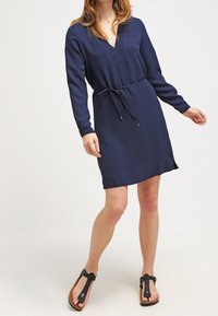 Vestido azul marino con cuello en v, mangas largas y lazo en la cintura. Tejido suave con ajuste relajado y aberturas laterales. Lleva sandalias marrones.