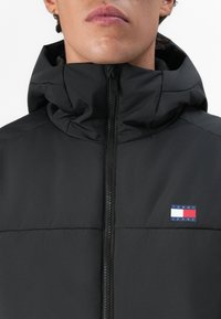 Vīrietis ģērbts melnā pūkainā jakā ar augstu apkakli, ko var aizvērt, ar nelielu Tommy Jeans logo uz krūšu.