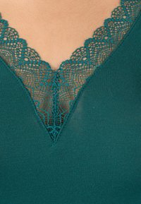 Top turchese con scollatura a V profonda, ornato da un intricato bordo in pizzo a forma di scallop, con una superficie liscia a costine e tessuto morbido.