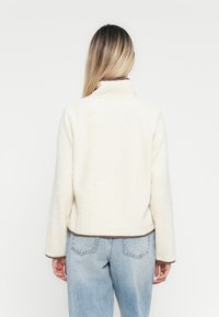 Crème-kleurige fleece jas met een kraag, voorzien van een bruine zoom en manchetten. Ontworpen met een relaxte pasvorm en een pluizige textuur.