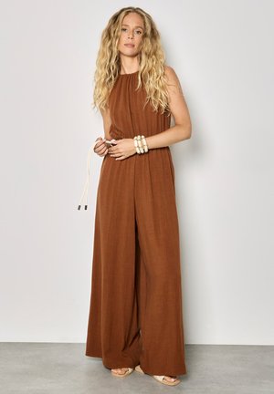 Kobieta o długich kręconych blond włosach, ubrana w brązowy, szerokodostępny kombinezon bez rękawów, trzymająca biały sznurkowy pas, stojąca na tle gładkiej ściany.