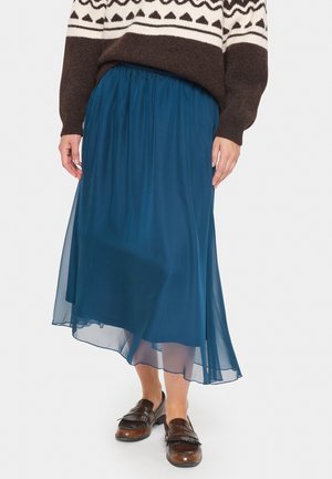 Persoon draagt een bruin gebreide trui met patroon, een blauwe midi-rok met transparante overlay en bruine leren loafers, staand op een witte achtergrond.