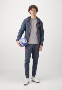 Sporty outfit med en navy jakke med orange detaljer, grå skjorte og navy bukser. Holder en farverig fodbold. Hvite fodboldstøvler.