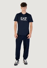 Camiseta de algodón azul marino con el logo blanco de EA7, combinada con pantalones de chándal a juego en azul marino. Las zapatillas presentan colores negro, gris y blanco.