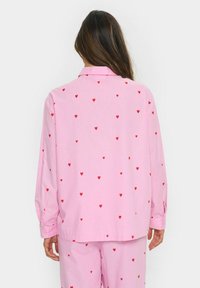 Camicia a maniche lunghe rosa con piccoli motivi a forma di cuore rossi. Il tessuto sembra liscio con un colletto classico e polsini con bottoni.