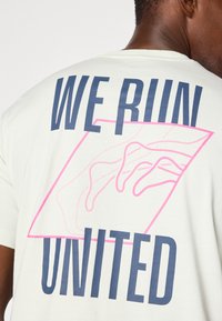 Tricou de culoare deschisă, cu text îndrăzneț în nuanță de navy "WE RUN UNITED" și un contur roz al unei mâini într-o formă geometrică.