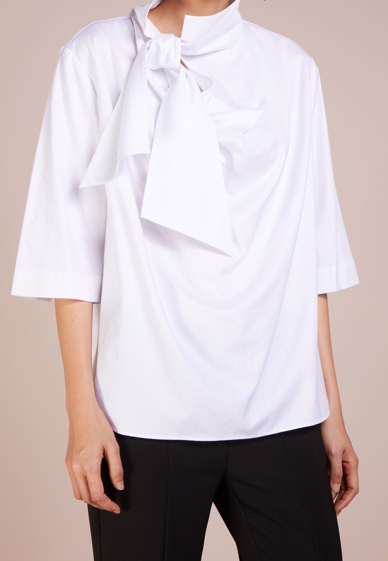 Femme portant un chemisier blanc avec un grand nœud au col et des manches jusqu'au coude, associé à un pantalon noir.