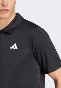 Crna polo majica od glatkog, elastičnog materijala s trostrukim bijelim logotipom na lijevoj strani grudi i otvorom s dvije dugmadi.