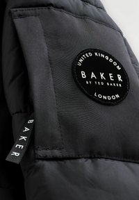 Melnā mīkstā jaka ar apļa izšuvuma uzlīmi ar tekstu: "BAKER by TED BAKER" un papildus auduma etiķeti ar vārdu "BAKER".