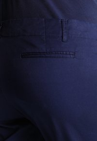 Mörkblå bomullsbyxor med en slät textur, som har en bakficka med diskret sömnadsdetalj och en dragkedja.