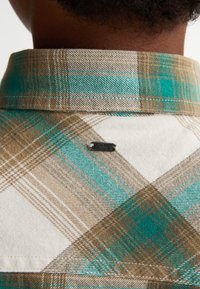 Karo košile v odstínech tyrkysové a hnědé, s límečkem na knoflíky. Na zadní straně se nachází kovový štítek s logem. Viditelná bavlněná textura.