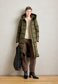 Abrigo largo y voluminoso de color verde oliva con capucha, puños acanalados y cremallera frontal. Llevado sobre un suéter beige y pantalones marrones, combinado con botas de tacón negras.