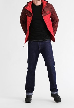 Rote und braune gesteppte Jacke mit Reißverschluss, schwarzes Hemd, dunkelblaue Jeans und schwarze Schuhe. Die Jacke verfügt über eine Kapuze und Brusttaschen.