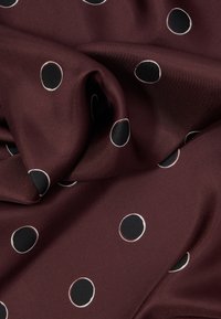 Μπορντό ύφασμα με λεία υφή, που παρουσιάζει ένα σχέδιο από μαύρους κύκλους με λευκό περίγραμμα, διάσπαρτους στην επιφάνεια.