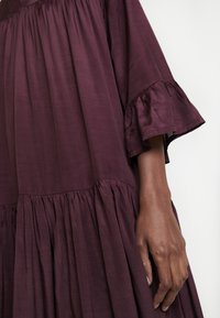 Robe violet foncé avec tissu froncé et volanté, manches évasées, montrée en gros plan sur le bras et la main d'une personne sur le côté.