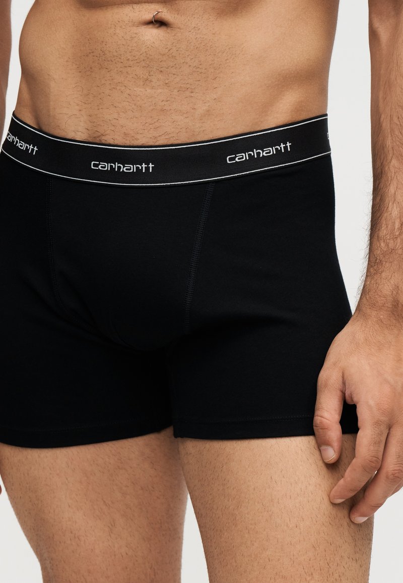 Männlicher Torso mit schwarzen Carhartt Boxershorts mit gebrandetem Bund, zeigt Bauchnabelpiercing und einen Teil der rechten Hand, die am Oberschenkel ruht.