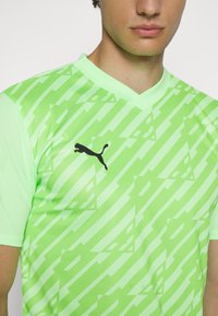 Zaļš, īsām piedurknēm sporta krekls ar V veida kakla izgriezumu. Ar �ģeometrisku rakstu un melnu Puma logo uz krūškurvja. Izgatavots no elpojoša auduma.