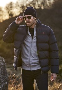 Svart dunjakke med horisontal søm, grå glidelåslaget innerlag, denim jeans og mørk lue; iført solbriller; ute.